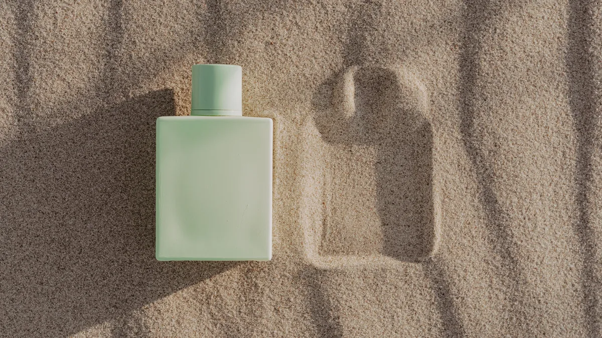 A light green bottle of perfume on a sandy surface, with the bottle's shadow cast to the left.