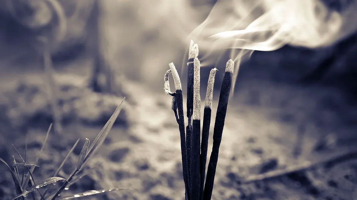 A cluster of incense sticks with smoke rising from them, set against a blurred background that appears to be a sandy surface.