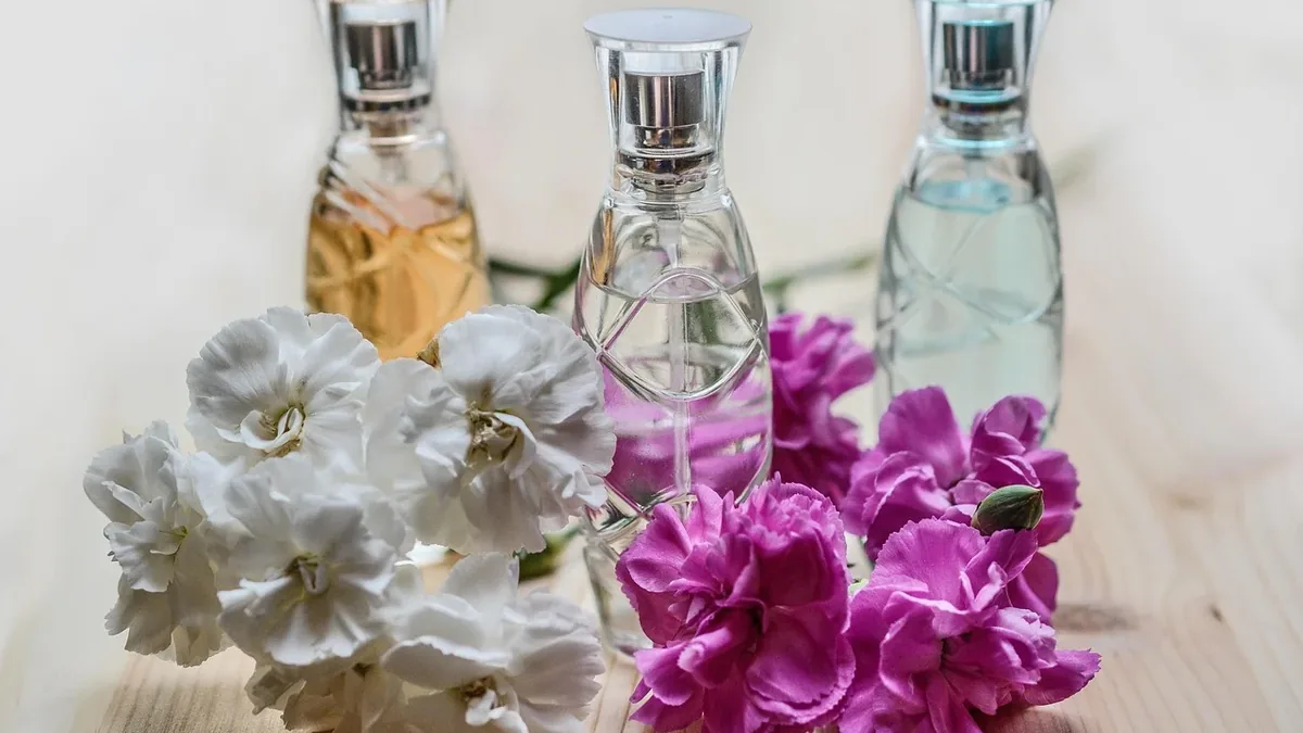 Three glass bottles of perfume on a light-colored wooden surface, with white and pink flowers in front of them.